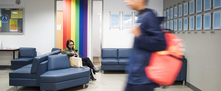 A smiling girl is sitting comfortably in the waiting area in front of the Centre for Health Sciences Faculty and Administration reception and looking at her phone.