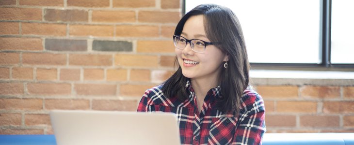 Female student is sitting casually in the campus lounge and working on her laptop while she is looking away and smiling.