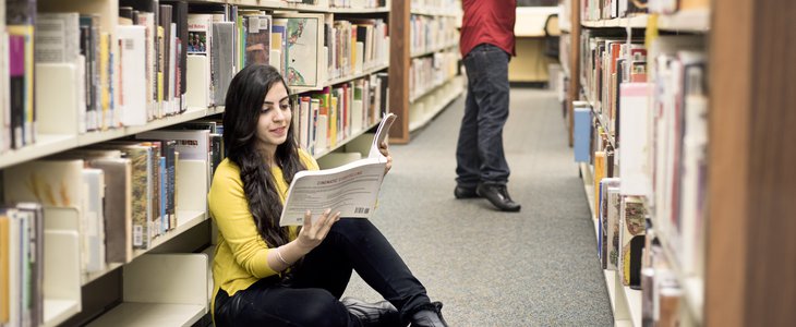 A female student is sitting on the floor of the library and reading a book and smiling. In the background there is a man pulling out a book from the shelves.
